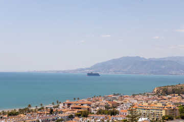 Obraz premium Coastal view of vibrant seaside town with colorful buildings, palm trees lining shore, large cargo ship sailing in the clear blue waters under a bright summer sky, capturing essence of summer in Spain