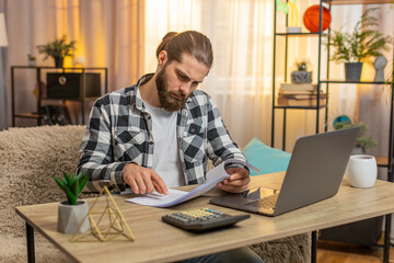 Young serious man checks papers at home table reading fine print new agreement with deep concern...