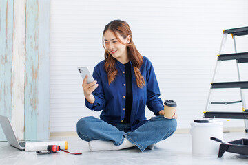 Asian young beautiful woman long curly hair wearing blue shirt jeans sit on floor using smartphone consult with friend short break after painting wall around rollers brush ladder renovate room concept