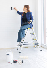 Asian young beautiful woman long curly hair wearing blue shirt jeans sitting on ladder holding  paintbrush preparing repairing room painting wall empty house or apartment renovation decorating concept