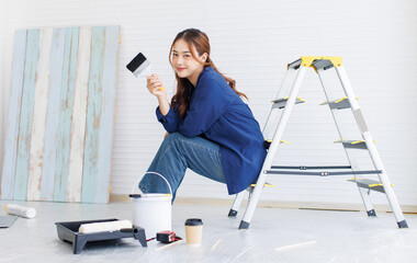 Asian young beautiful woman long curly hair wearing blue shirt jeans posing sitting back in ladder look at camera while repairing room around rollers brushes paint bucket renovate decorating the house