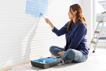 Asian young beautiful woman smiling cheerful long curly hair wearing blue shirt jeans sitting on floor painting wall with roller brush light blue for new home repair renovation decoration concept.