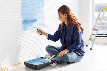 Asian young beautiful woman smiling cheerful long curly hair wearing blue shirt jeans sitting on floor painting wall with roller brush light blue for new home repair renovation decoration concept.