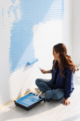 Asian young beautiful woman smiling cheerful long curly hair wearing blue shirt jeans sitting on floor painting wall with roller brush light blue for new home repair renovation decoration concept.