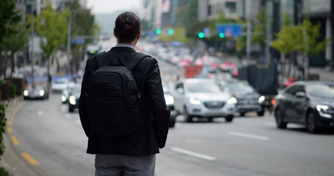 Man in smart casual suit with backpack stands with back to camera, gazing at slow-moving traffic on congested Gangnam street during peak hour, might be waiting for someone. Selective focus shot