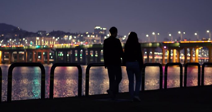 Silhouetted tourist couple walking hand in hand along Han River at late hour, blurred city lights and glowing Hannam Bridge in background. Nighttime stroll as young man and woman share quiet moments