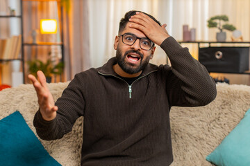 Indian man at home sits on sofa and covers face with hand in frustration after realizing mistake....