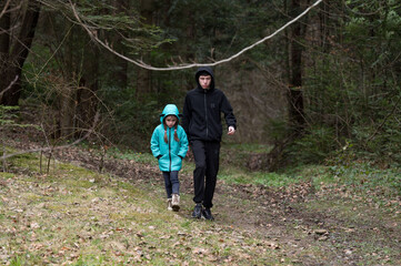 Two children explore a tranquil forest path on a chilly day, surrounded by lush greenery and fallen leaves, creating a moment of connection and adventure in nature