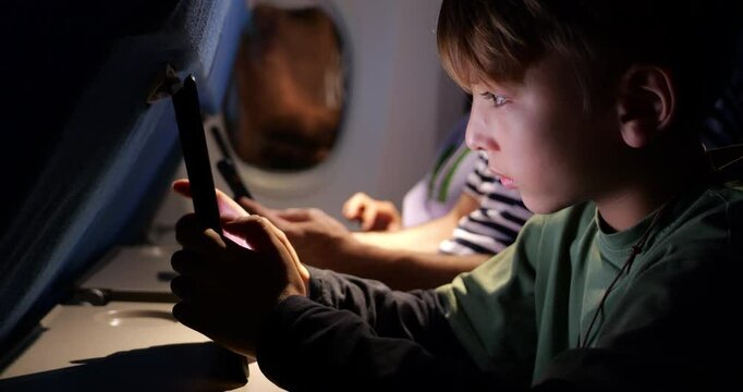 Young passenger engaged with tablet during nighttime flight, appearing to solve puzzle game. Boy seated beside parent also immersed in digital devices, reflecting modern family travel