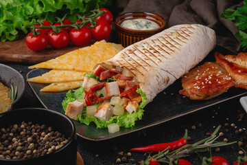 Close-up of grilled chicken shawarma wrap filled with cheese cubes, pineapple, lettuce, and tomatoes, served on a black plate with herbs and condiments