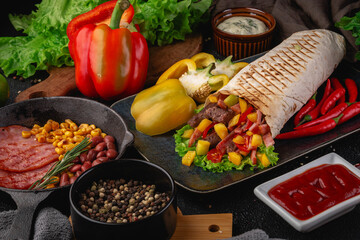 Delicious tortilla wrap filled with grilled beef, sweet corn, bell peppers, lettuce, and cherry tomatoes, surrounded by fresh vegetables and condiments on a rustic black background