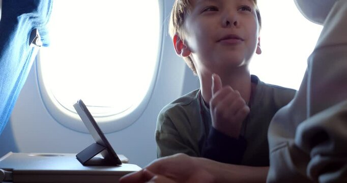 Young boy using tablet during daytime flight, actively discussing something with his mother seated next to him in economy class cabin. He taps screen to show her, then turns to speak to mother