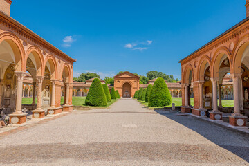 The 15th-century Charterhouse of Ferrara, set against a lush green lawn with San Cristoforo church. Ancient cemetery cloister featuring hanging lamps, tombs, and sculptures in Ferrara city of Italy