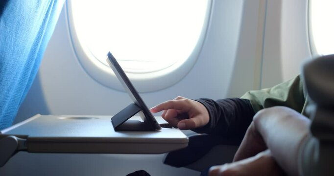 Young passenger boy tapping and scrolling through digital content on tablet resting on airplane tray table by window, utilizing in-flight wireless technology for entertainment - Powered by Adobe