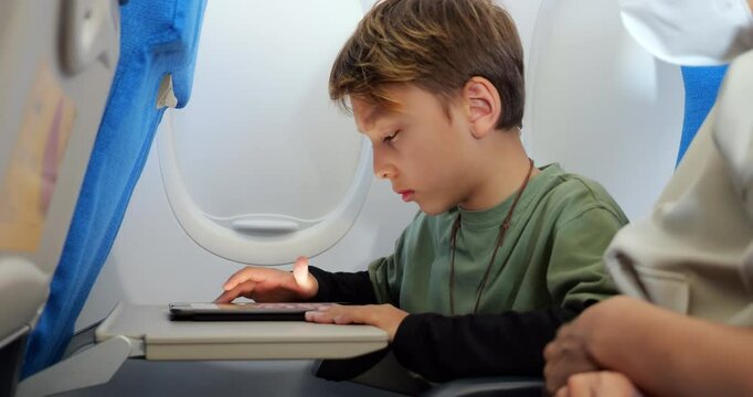 Passenger boy appearing puzzled while interacting with unfamiliar application on his tablet, tapping screen with uncertain expression as his mother, seated beside him, tries to help.
