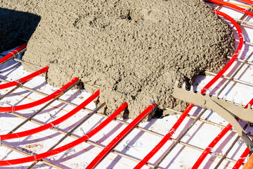 Concrete pouring over underfloor heating pipes and mesh, Red underfloor heating pipes attached to a steel mesh grid being covered with freshly poured concrete.