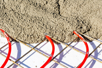 Concrete pouring over underfloor heating pipes and mesh, Red underfloor heating pipes attached to a steel mesh grid being covered with freshly poured concrete.