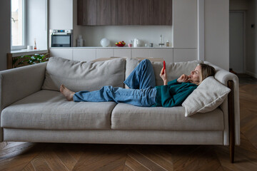 Relaxed midlife woman rest on couch focused looking on screen phone, reading news, browsing internet web. Interested pleased female lying on cozy sofa online shopping with smartphone ease from home.