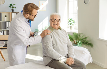 Obraz premium Senior happy man patient in medical clinic talking with male young friendly doctor wearing stethoscope sitting on couch in office during examination. Doctor support and health care concept.