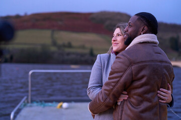 Fototapeta premium Smiling couple in warm clothing embracing on pier at dusk
