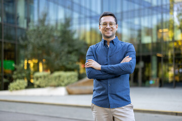 A confident businessman, smiling with arms crossed, stands confidently in front of a modern office building on a sunny day.