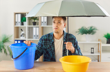 Man troubled at home holding umbrella among plastic bucket and basin, hole, crack in ceiling, water...