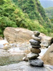 A carefully balanced stack of stones stands in the foreground, symbolizing harmony and patience, while lush green mountains and smooth river rocks create a peaceful backdrop.