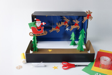 Handmade Christmas diorama in cardboard box featuring Santa Claus in sleigh with reindeer. DIY paper craft scene with colored paper. Winter holiday and Christmas celebration concept. Front view
