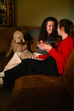 Lesbian couple having cocktails and sitting on sofa with dog at home