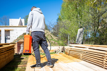 Fototapeta premium A worker carries an orange sprayer at a construction site. He stands on wooden planks with stacks of lumber nearby. A partially built structure and green trees are visible under a clear blue sky