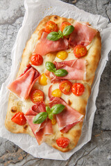 Pinsa Romana with with cherry tomatoes, ham, mozzarella cheese and basil closeup on parchment paper on the table. Vertical top view from above