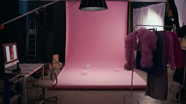 No people zoom out shot of professional photo studio interior with seamless pink background paper, lighting equipment, photographer workstation with PC and stylish clothes rail - Powered by Adobe