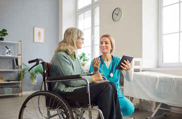 Smiling female nurse in blue uniform kneeling beside old woman in wheelchair, discussing treatment and test results. Friendly healthcare worker talking to disabled senior patient in medical office.