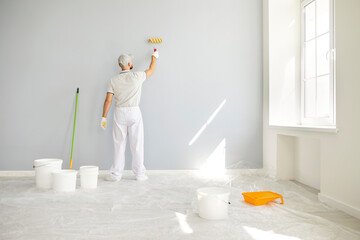 Back view of male painter in white uniform clothes painting light grey wall with paint roller....