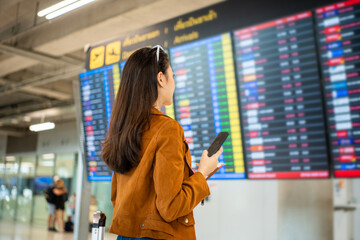happy young asian woman traveler holding boarding pass ticket, passport and smartphone at the airport terminal with her luggage and smiling at camera, cheerful tourist female having holiday trip.