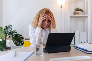 Stressed businesswoman holding head in hands while working on laptop at desk in modern office, facing work pressure or burnout.