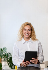 Fototapeta premium Smiling businesswoman holding a tablet in a bright modern office. Confident and approachable, she stands in a professional environment with plants and minimal decor.