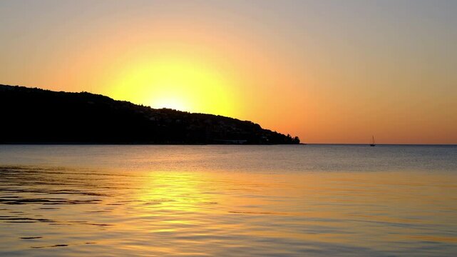 Tranquil seascape at sunset with golden reflections on the calm water and the sun setting behind a silhouette of a coastal hill.