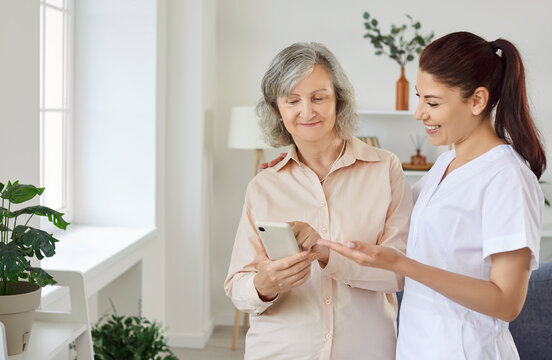 Young friendly smiling female nurse showing elderly woman how to use smartphone. Caregiver teaching senior person to handle with mobile phone. Nursing home service and patient support concept.