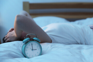 Frustrated young woman suffering from insomnia on bed indoors, focus on alarm clock