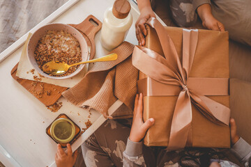 Children in pajamas holding a wrapped gift while enjoying breakfast in bed with oatmeal, milk, candle, and cozy textures. A warm family moment in soft morning light