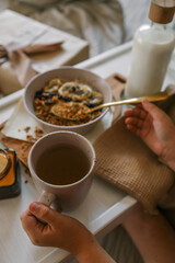 Child’s hands holding spoon and mug while enjoying breakfast in bed with granola, milk, candle, and a wrapped gift in the background. Cozy and calm morning scene