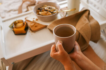 Child’s hands holding spoon and mug while enjoying breakfast in bed with granola, milk, candle, and a wrapped gift in the background. Cozy and calm morning scene