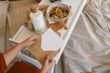 Child opening kraft envelope with card on a cozy bed, next to breakfast tray with granola and milk,...