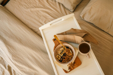 Flatlay of cozy breakfast tray with granola, milk, amber candle, and napkin on a soft beige bed....