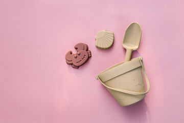 Flat lay of silicone beach toys including crab and shell molds with a beige bucket and shovel on pastel pink background. Modern eco-friendly children's play set.