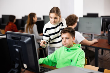 Interested teen schoolgirl talking to diligent focused classmate sitting at computer in classroom during informatics lesson..