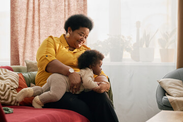 Happy African American grandmother holding little grandchild while playing with baby on cozy couch in living room