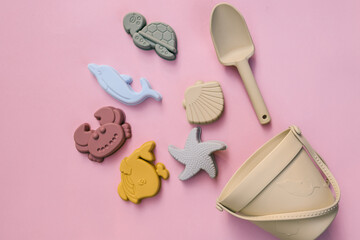 Set of pastel silicone beach toys arranged on pink background, including dolphin, crab, turtle, starfish, fish, seashell, bucket, and shovel in minimalistic eco-friendly style