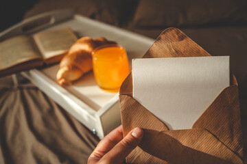 Close-up of a hand holding a kraft envelope with a blank card, with a breakfast tray in the...